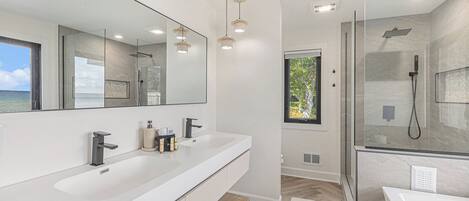 A modern bathroom with a large mirror, two sinks, a shower, and a view of the sea.