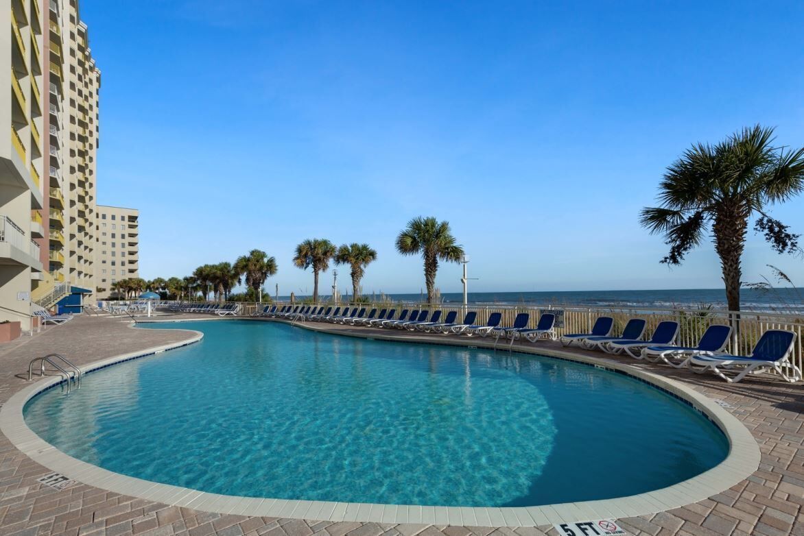 A swimming pool with lounge chairs, palm trees, and a building in the background.