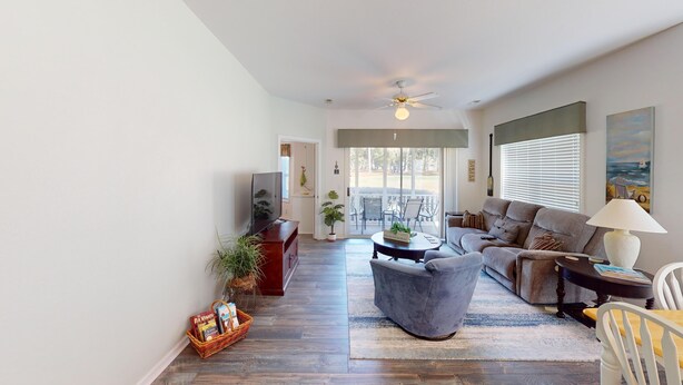 A spacious living room with a grey sofa, a coffee table, and a TV. There is a bookshelf, a plant, and a window with blinds. - River Creek 2 Unit 2002 (Sunset Beach)