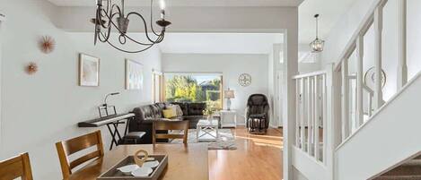 A spacious living area with a wooden dining table, a sofa, and a baby rocker. A chandelier hangs from the ceiling, and there are framed pictures on the walls.