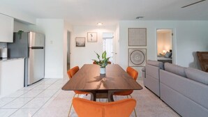 A modern kitchen with a stainless steel refrigerator, a wooden dining table with orange chairs, and a grey sofa in the living area. - Fillmore Unit 11 (Cape Canaveral)