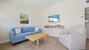 Una sala de estar con un sofá azul, un sillón blanco, una mesa de centro de madera, un televisor montado en la pared y un ventanal con vistas a árboles.