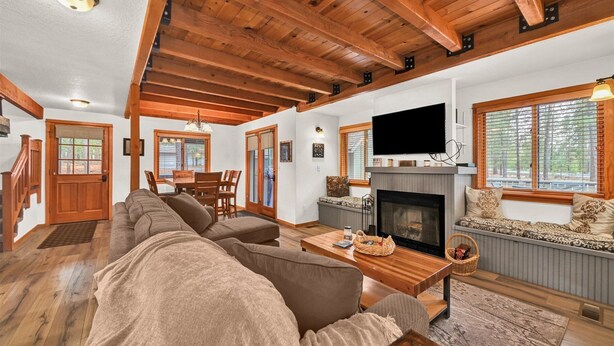 A spacious living room with a fireplace, wooden ceiling beams, a television mounted above the fireplace, and a large window with blinds. - Otter Hot Tub Retreat Near Sharc (Bend)