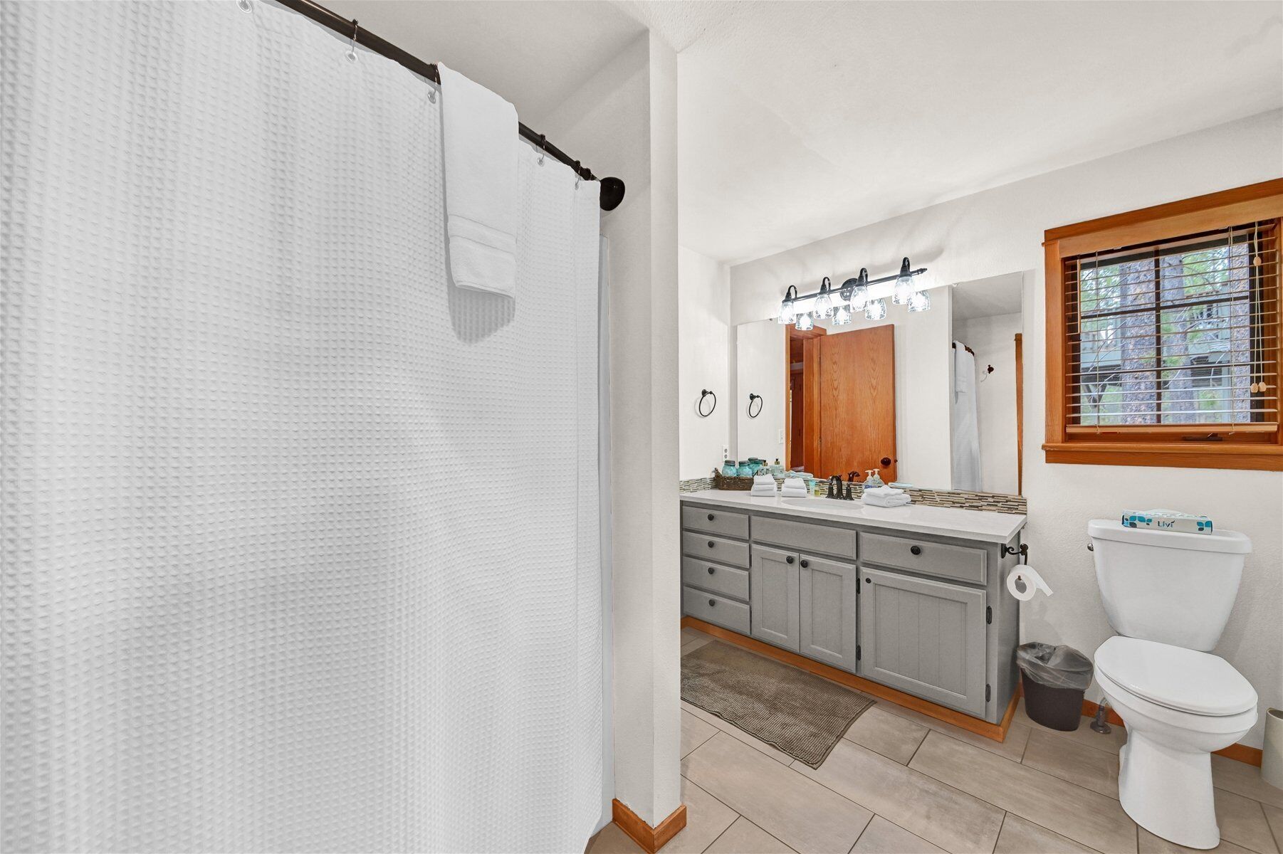 Ein Badezimmer mit Duschvorhang, Waschtisch mit Spiegel, Toilette und einem Fenster mit Jalousien.
