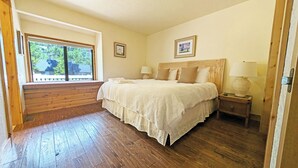 A bedroom with a large window, wooden flooring, a bed with white bedding, two bedside tables with lamps, and two framed pictures on the wall. - East Butte 14 (Bend)