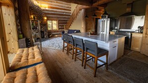 Een gezellig houten blokhut interieur met een stenen openhaard, een houten bank, een keuken met een groot eiland en barstoelen, en een trap naar een vide.