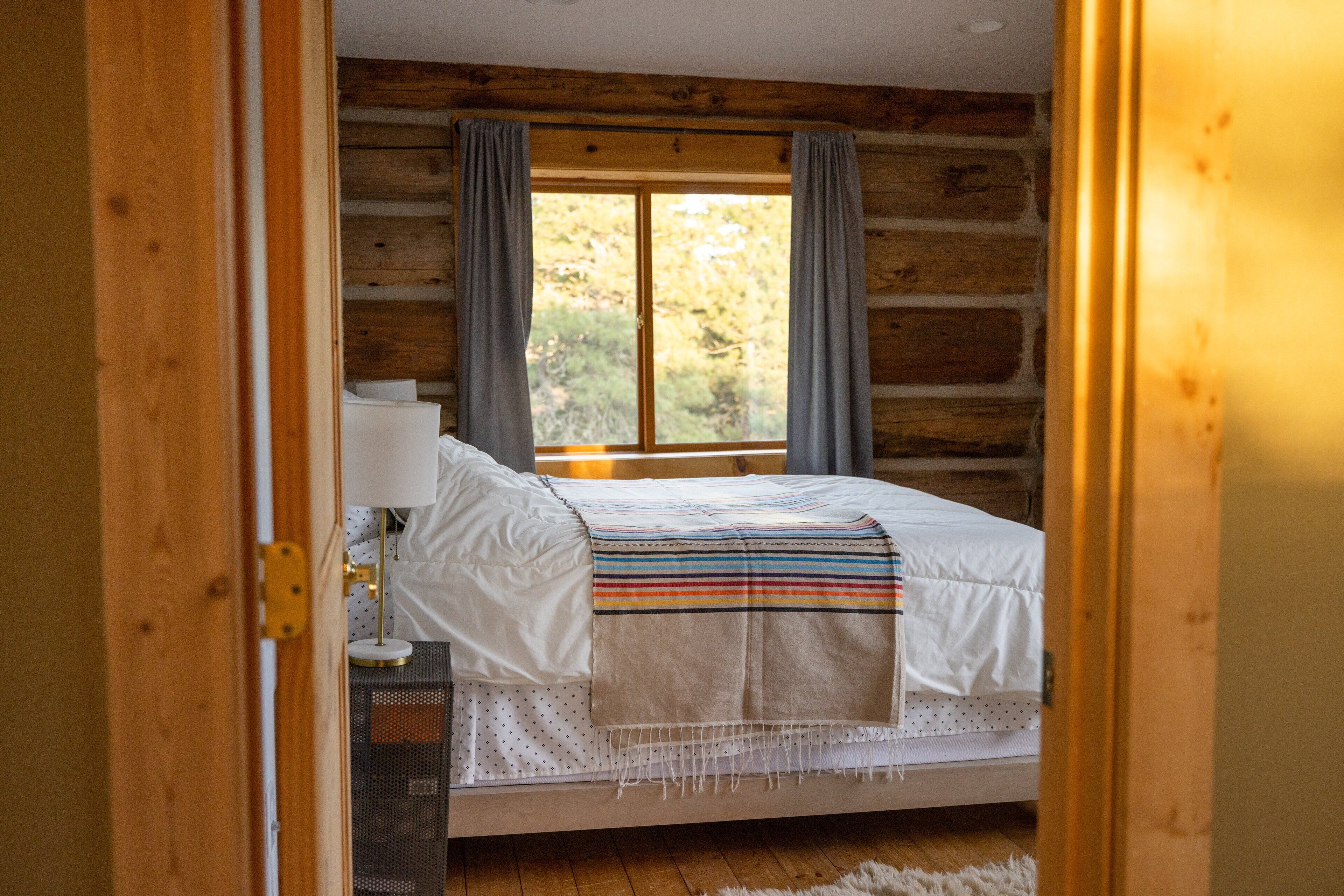 Un dormitorio de cabaña de troncos con una cama, una mesita de noche, una lámpara y una ventana con cortinas.