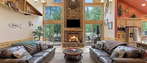 Un salón amplio con sofás de cuero, una chimenea de piedra y grandes ventanales con vistas a árboles.