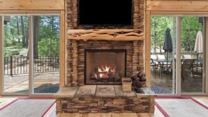 Ein Kamin aus Stein mit einem Fernseher darüber, ein Feuer brennt und ein Blick ins Freie durch große Glastüren.