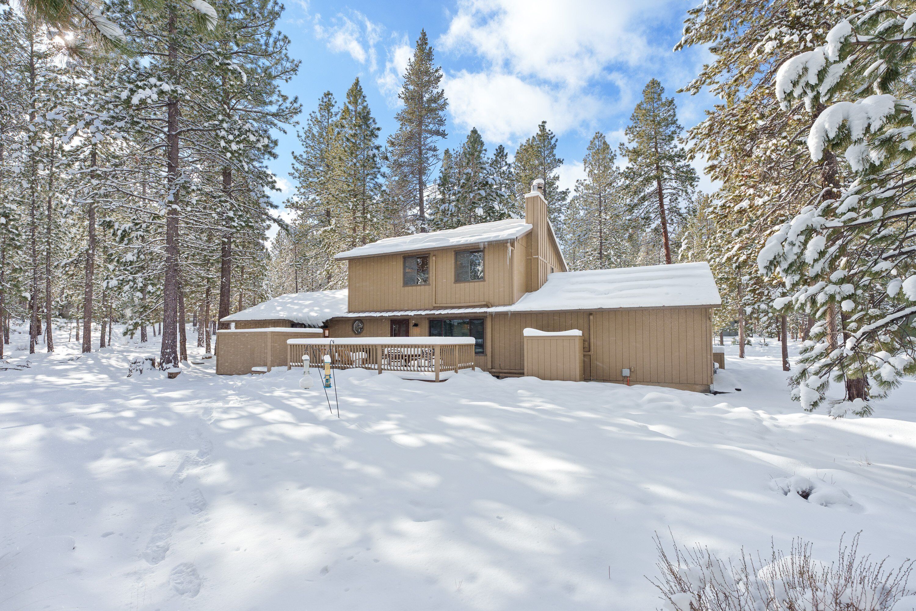 A wooden cabin with a large deck and a chimney, surrounded by snow-covered trees.