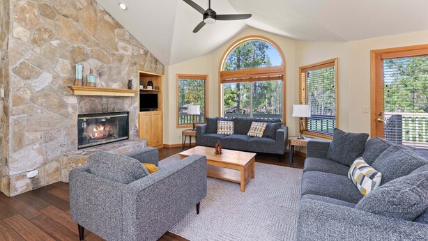 A living room with a stone fireplace, a ceiling fan, and a large window. - Sunriver Golf Views Hot Tub Escape (Bend)
