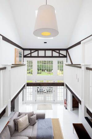 A spacious room with a large bed, a hanging light fixture, and a view of the outdoors through large windows. - Bridgehaven Estate by Rove - Luxe Home With Pool (Bridgehampton)