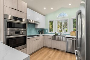 A modern kitchen with stainless steel appliances, light wood cabinets, and a view of greenery outside. - Marbella Lane Wave HS w EV Charger, Oceanfront! (Oak Harbor)