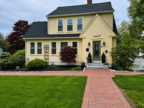 A two-story yellow house with a brick pathway leading to the front door. - Classic New England Retreat in the Heart of Boothbay Harbor (Boothbay Harbor)