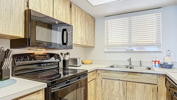 A modern kitchen with wooden cabinets, a stainless steel stove, a microwave, and a window with blinds. - Oceania Beach Club 203 (New Smyrna Beach)