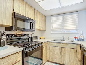 A modern kitchen with wooden cabinets, a stainless steel stove, a microwave, and a window with blinds. - Oceania Beach Club 203 (New Smyrna Beach)
