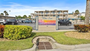 A hotel entrance with a sign reading "Win-San" and the number 111. There are cars parked in the lot, and a grassy area with bushes and a palm tree. - Win-san C102 (New Smyrna Beach)