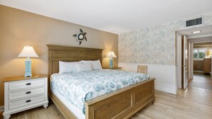A bedroom with a wooden bed, two bedside tables with lamps, a wall clock, and a door leading to another room. - Win-san C102 (New Smyrna Beach)