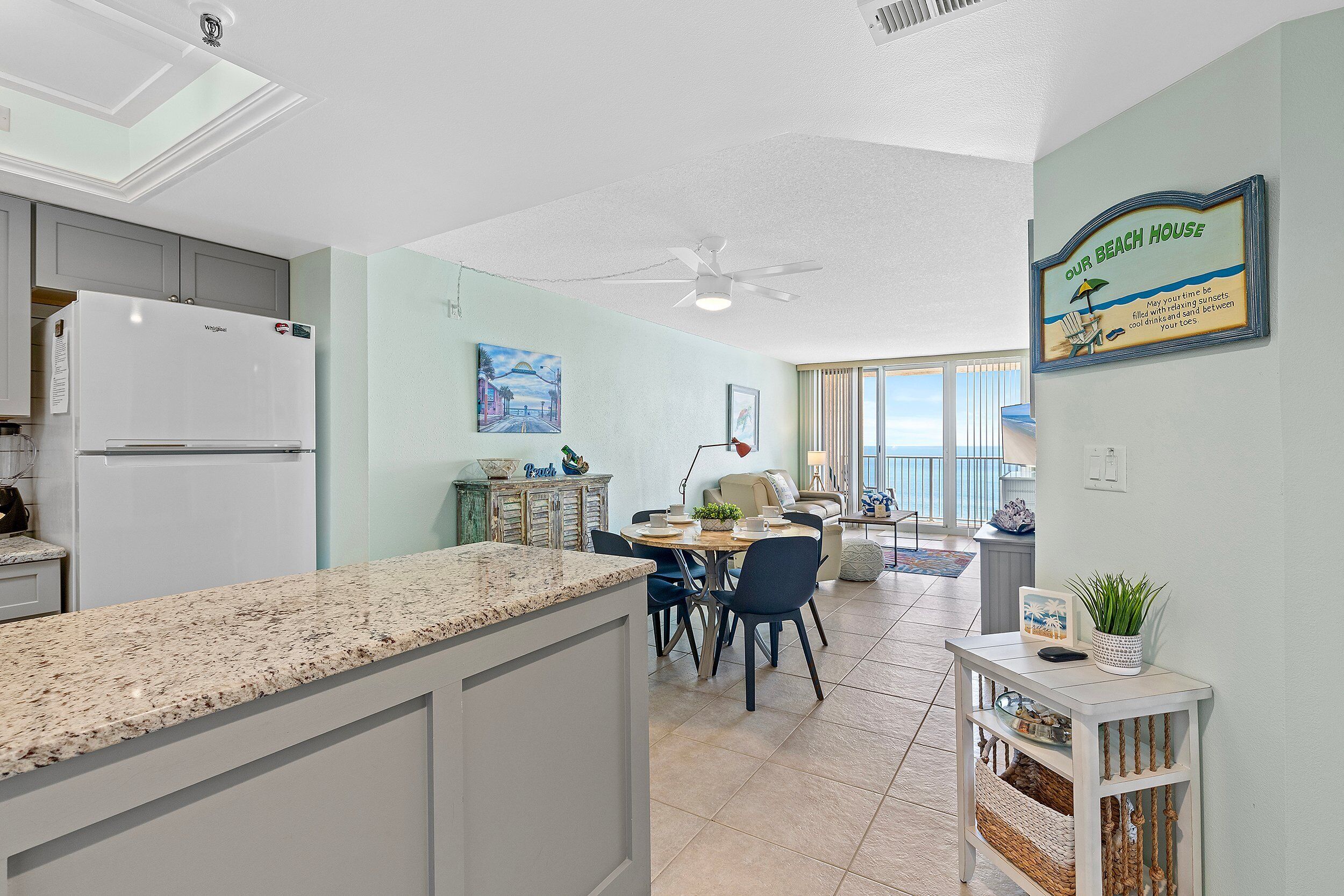 A modern kitchen with a built-in refrigerator, a dining area with a round table and chairs, and a living space with a ceiling fan and a beach-themed sign.