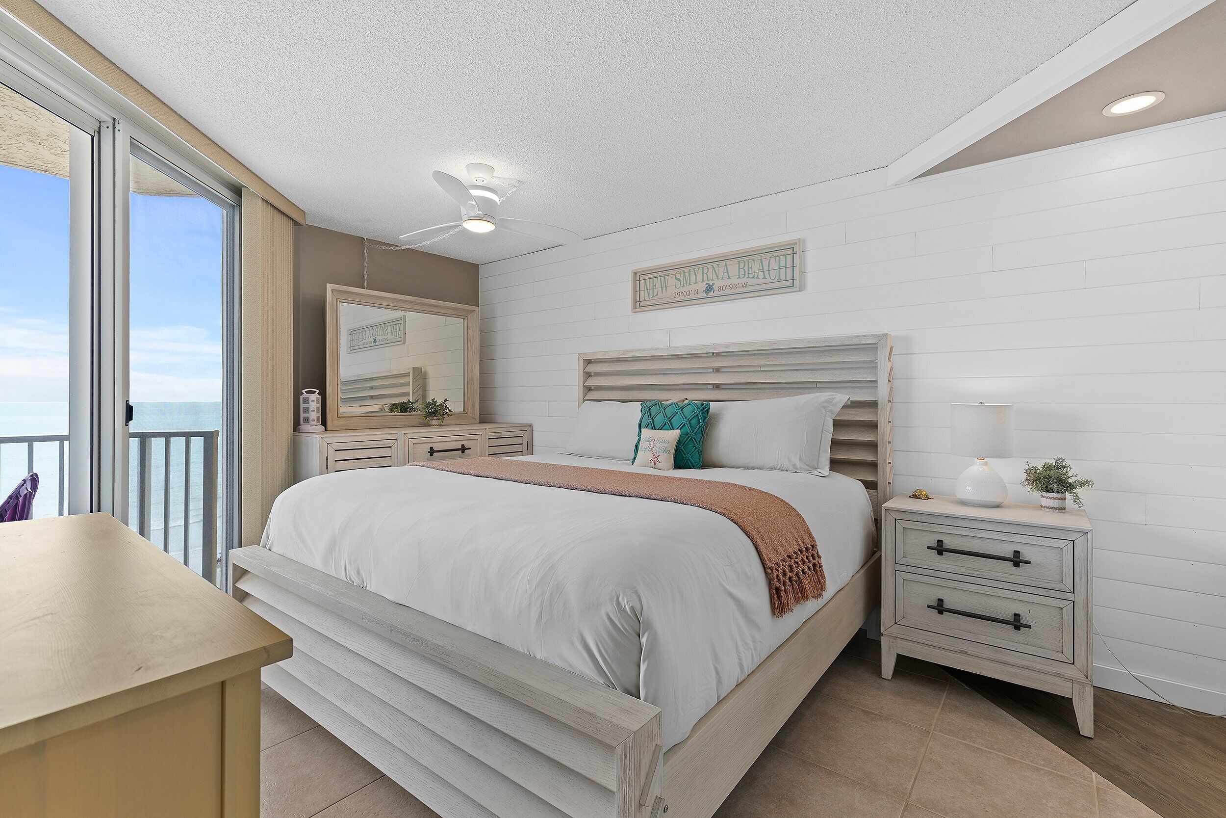 A bedroom with a bed, bedside tables, a nightstand, a ceiling fan, and a view of the beach.