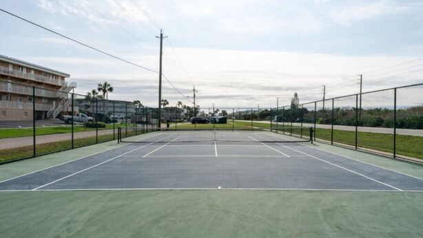 An outdoor tennis court with a green surface, surrounded by a fence, and adjacent to a building with multiple floors. - Ocean Club North C104 (New Smyrna Beach)