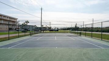 An outdoor tennis court with a green surface, surrounded by a fence, and adjacent to a building with multiple floors.