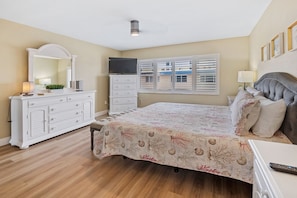 Un dormitorio con cama, cómoda, espejo, televisor y ventana con persianas.