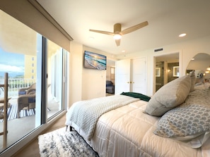 A bedroom with a large bed, a ceiling fan, a painting, and a view of the outside. - Oceania Beach Club 201 (New Smyrna Beach)