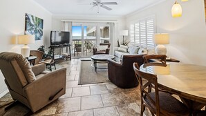A spacious living room with a flat-screen TV, comfortable seating, a dining table, and a balcony with a view.