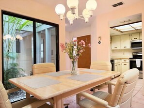 Zona pranzo con tavolo in legno, sedie color beige, un vaso con fiori e vista sull'esterno tramite una porta in vetro.
