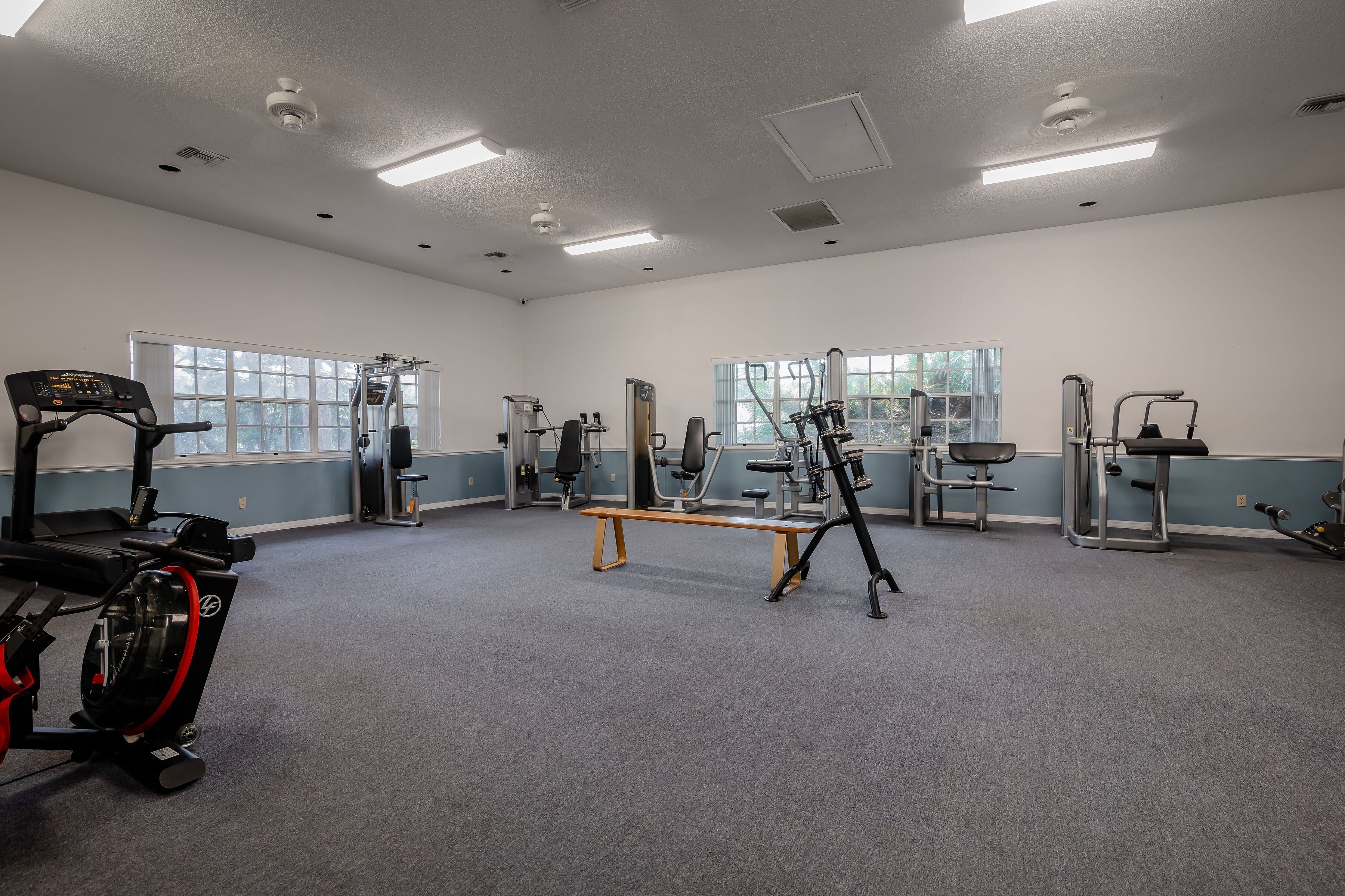 Ein großzügiger Fitnessraum mit unterschiedlichen Trainingsgeräten, großen Fenstern und einer neutralen Farbgestaltung.