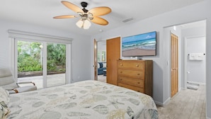 Une chambre à coucher comprenant un lit, un ventilateur de plafond, un téléviseur fixé au mur, une commode en bois et une porte-fenêtre coulissante donnant sur un balcon.