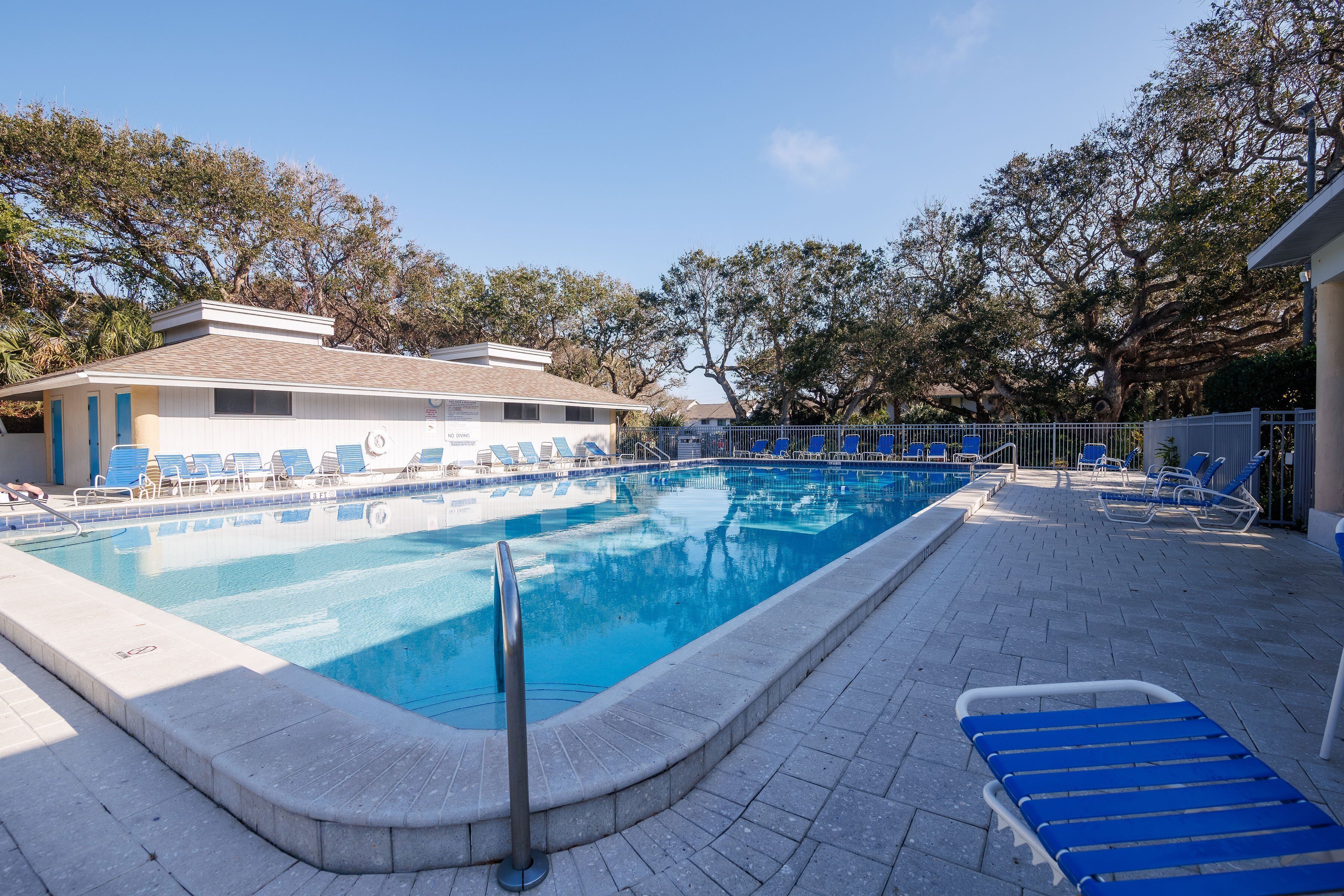 A swimming pool with a tiled deck, surrounded by lounge chairs and a building with a sloped roof.
