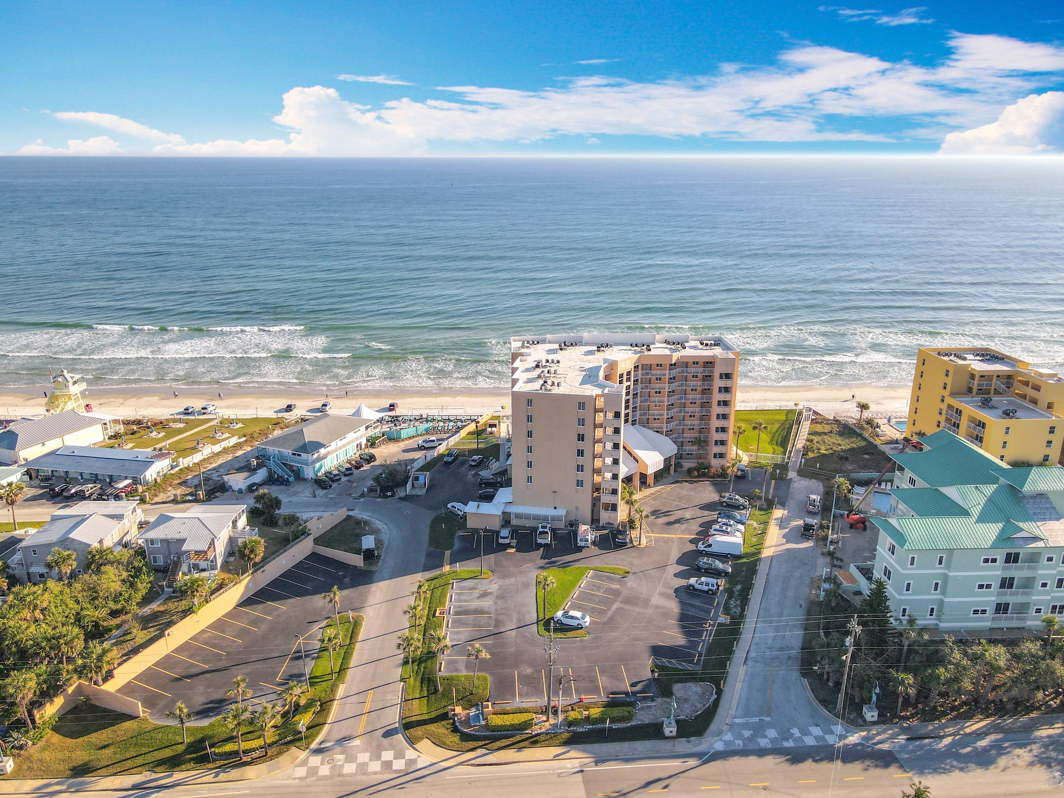 A coastal hotel with multiple buildings, a parking lot, and a beach view.