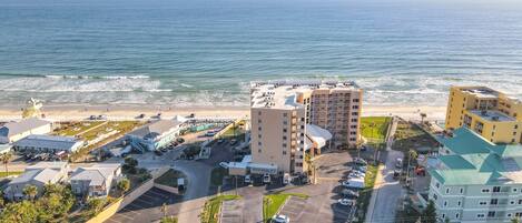 A coastal hotel with multiple buildings, a parking lot, and a beach view.