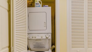 Ein kleiner Waschraum mit einer gestapelten Waschmaschine und einem Trockner, einer Flasche Purex Waschmittel und einer Tür mit Klappläden.