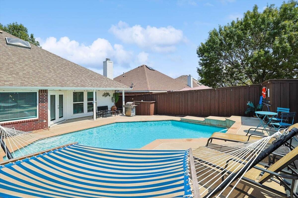 A backyard with a swimming pool, a covered patio, and outdoor furniture.