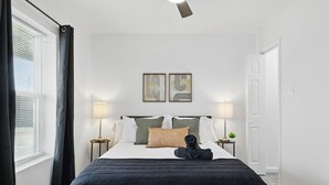 A neatly made bed with a black and white bedspread, two bedside lamps, and a window with curtains. - Modern 4BR 2BA Home With Hot Tub-grill & Huge Yard (Haltom City)