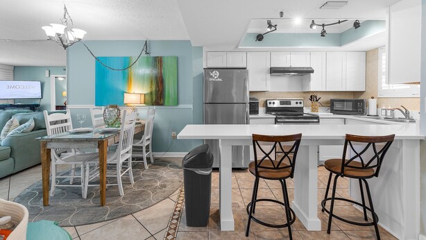 A modern kitchen with white cabinets, a stainless steel refrigerator, and a dining area with a glass table and white chairs. - Oceania Beach Club 803 (New Smyrna Beach)