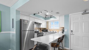 A modern kitchen with a stainless steel refrigerator, white cabinets, a large island, and a ceiling light fixture. - Oceania Beach Club 803 (New Smyrna Beach)