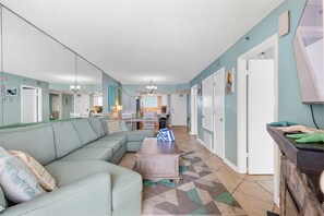 A spacious living room with a sectional sofa, a wooden coffee table, and a patterned rug. There are multiple doors leading to other rooms, and a kitchen area is visible in the background. - Oceania Beach Club 803 (New Smyrna Beach)