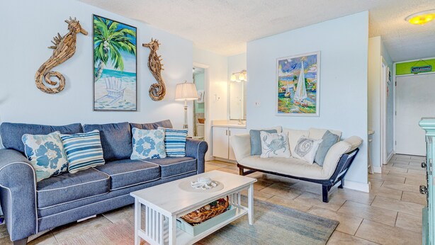 Ein Wohnzimmer mit einem blauen Sofa, einem weißen Couchtisch und zwei weißen Sesseln. An den Wänden befinden sich Kunstwerke, darunter ein Seepferdchen und ein Segelboot.