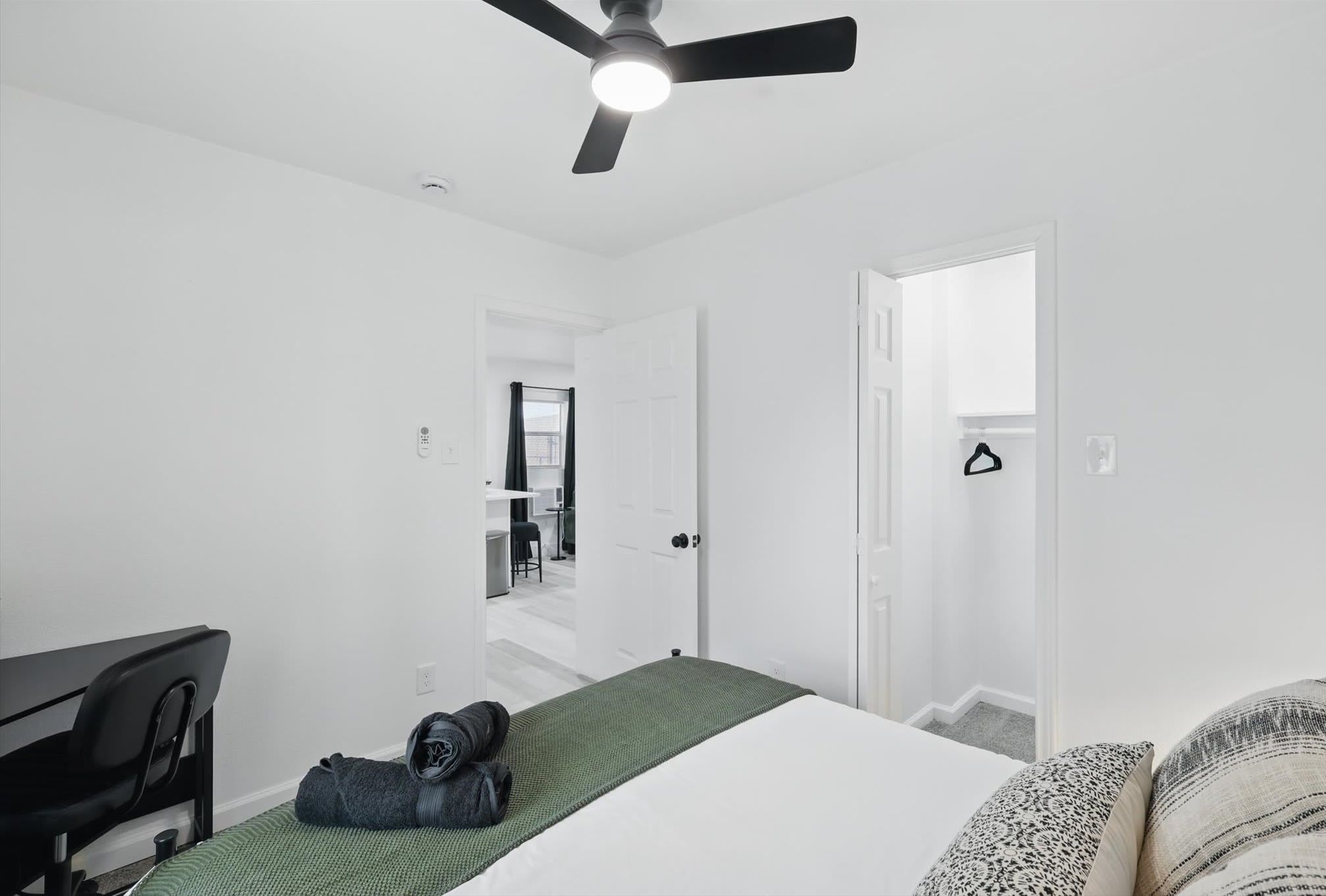 Quarto com cama, escrivaninha, cadeira, ventoinha no teto e porta que dá para uma escadaria.