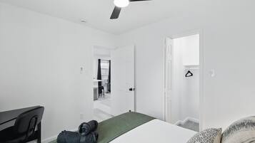 Quarto com cama, escrivaninha, cadeira, ventoinha no teto e porta que dá para uma escadaria.