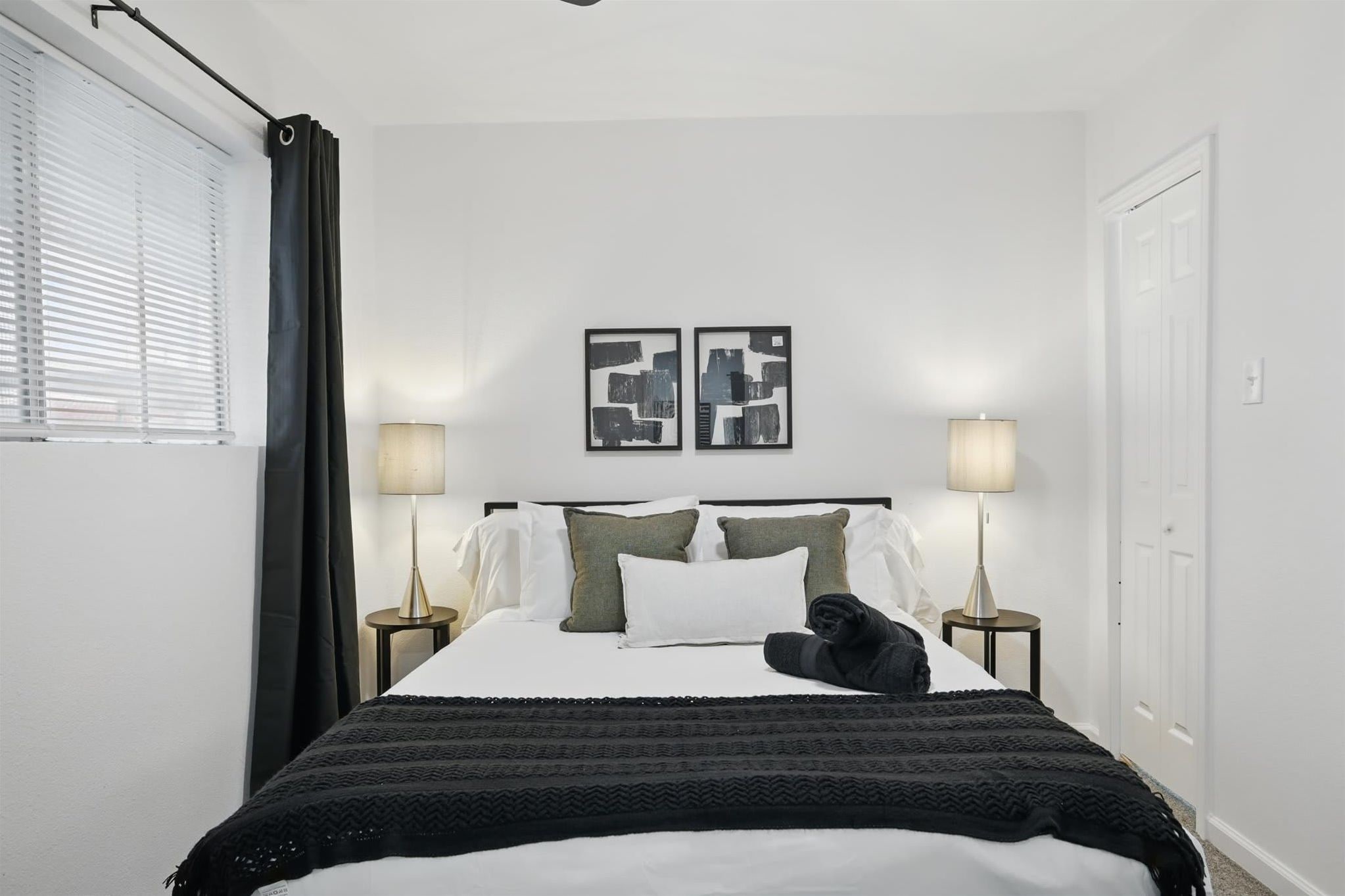 Une chambre bien rangée, avec un lit, deux lampes de chevet, une fenêtre avec des stores et une porte.