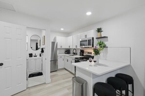 A modern kitchen with white cabinets, a stainless steel refrigerator, a microwave, and a coffee machine. There is a small island with a vase of flowers and wine glasses. - Modern 2BR Unit Hot Tub Outdoor Lounge (Haltom City)