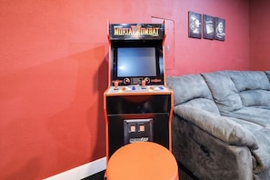En arkademaskine i retrostil med teksten 'Mortal Kombat', i et rum med rød væg og en grå sofa.