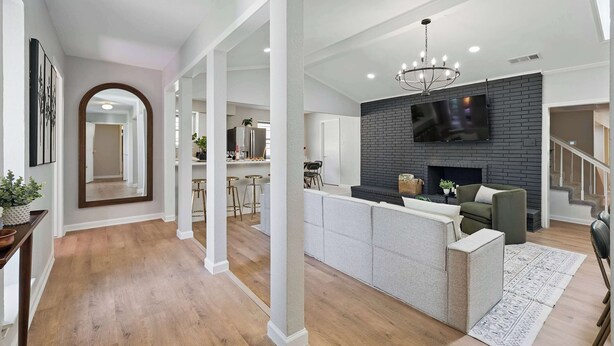 A spacious living area with a fireplace, a television mounted on a brick wall, and a modern kitchen in the background. - Spacious 5BR 3BA Retreat With Pool & Hot Tub (Duncanville)