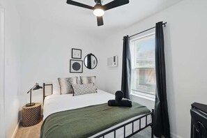 A bedroom with a bed, a ceiling fan, a window with curtains, a bedside lamp, and wall decorations. - Modern 2BR Unit 3 With Hot Tub (Haltom City)