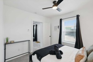 Un dormitorio con cama, ventilador de techo, ventana con cortinas, una puerta y una planta pequeña sobre una mesita consola.